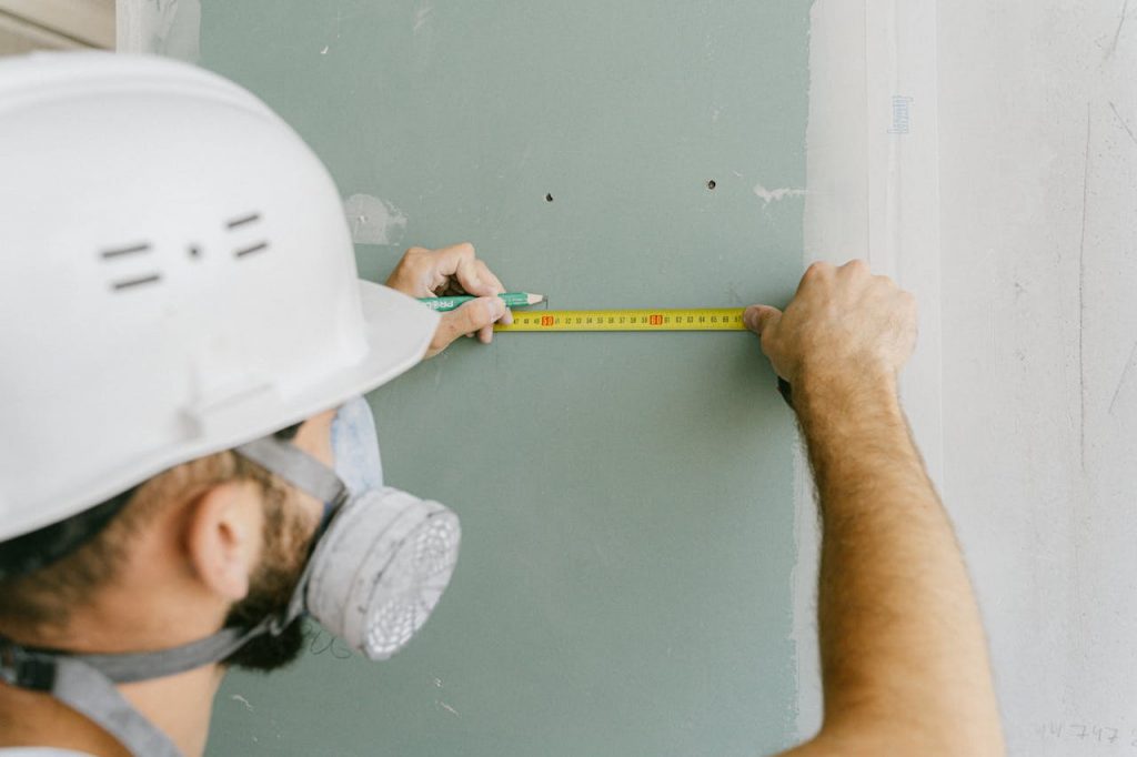pexels-photo-5493663 A construction worker precisely measuring drywall, wearing a helmet and mask for safety.